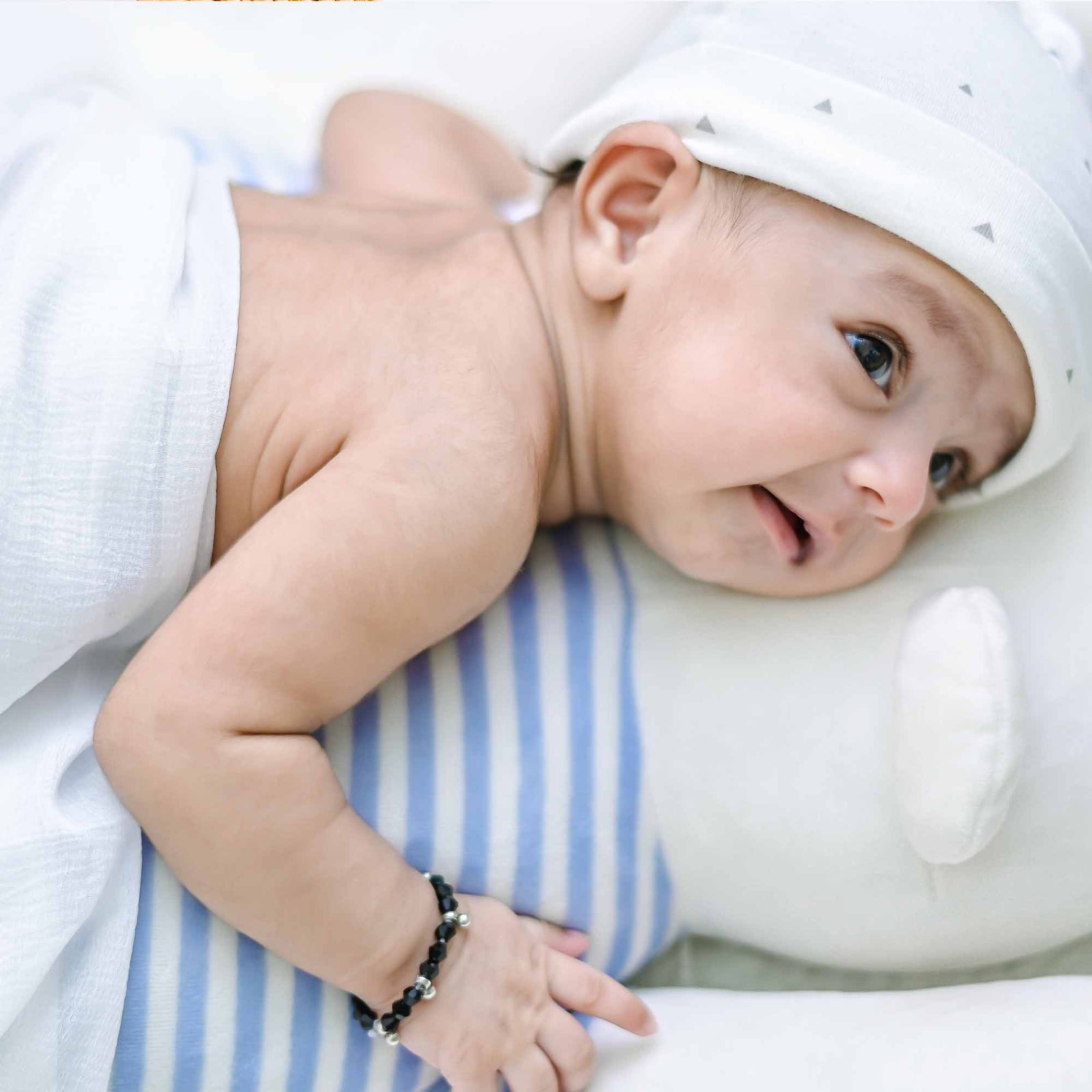 Newborn baby lying on a striped pillow, wearing a white hat with tiny triangles and a black beaded bracelet on their wrist.
