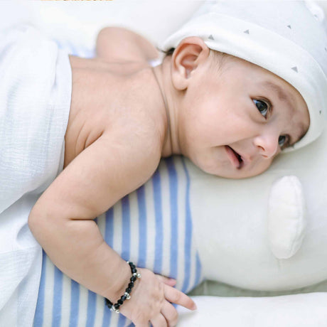Newborn baby lying on a striped pillow, wearing a white hat with tiny triangles and a black beaded bracelet on their wrist.