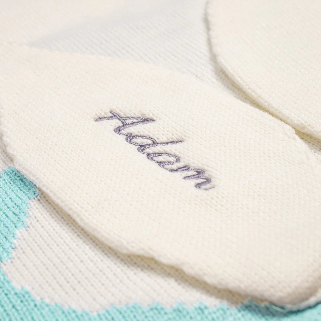 A close-up of a knitted blanket with the name "Adam" embroidered on it. The blanket is light-colored with a garter stitch border.