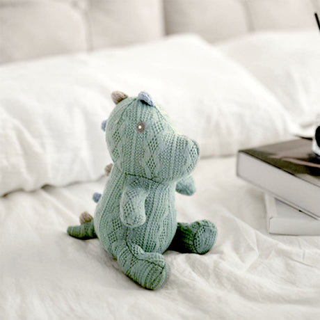 A charming knitted dinosaur toy in a soft seafoam green sits on a white bedspread, gazing out with tiny bead eyes. Its pose is playful and upright, suggesting curiosity and friendliness. The dinosaur is meticulously crafted with a textured knit pattern that gives it a cozy, hand-made feel, ideal for a child's gentle play or as a decorative nursery item.