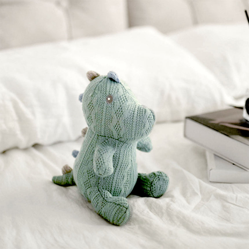 A charming knitted dinosaur toy in a soft seafoam green sits on a white bedspread, gazing out with tiny bead eyes. Its pose is playful and upright, suggesting curiosity and friendliness. The dinosaur is meticulously crafted with a textured knit pattern that gives it a cozy, hand-made feel, ideal for a child's gentle play or as a decorative nursery item.