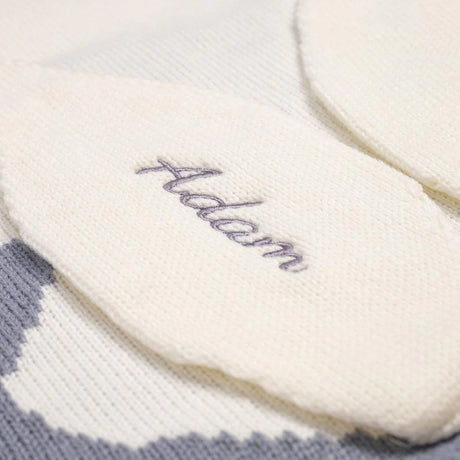 Close-up view of a white knitted baby blanket, showcasing the name 'Adam' elegantly embroidered in gray cursive script on the fabric, emphasizing the personalization detail and the soft texture of the blanket.