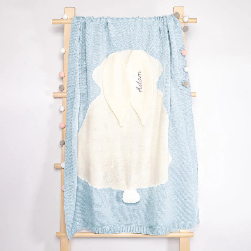 Personalized baby towel draped over a wooden rack, showcasing a delicate pastel blue color with a plush white hood featuring a playful pompom. The hood is elegantly embroidered with the name 'Adam’ in script, set against a clean and simple backdrop that highlights the towel's softness and inviting texture, ideal for a newborn's sensitive skin.