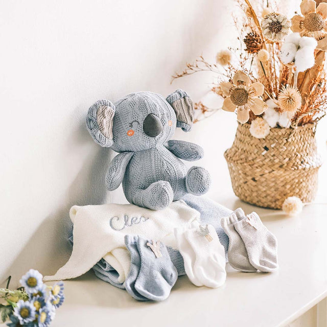 Charming baby gift set on a white shelf, featuring a hand-knitted gray koala plushie with orange button eyes and floppy ears, sitting next to a personalized white baby blanket embroidered with the name 'Cleo'. The set includes three pairs of soft baby socks in shades of gray and white, displayed in front of a rustic woven basket filled with dried flowers, creating a cozy and inviting nursery atmosphere.