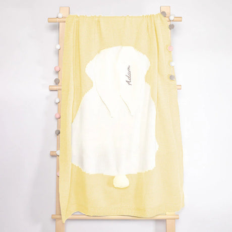A soft lemon yellow baby towel hangs on a wooden rack, adorned with a plush white hood featuring a cute pompom and elegantly embroidered with the name 'Adam’ in a graceful script. The towel's light, pastel shade contrasts beautifully against the subtle, natural tones of the wooden rack, which is decorated with small, round beads in soft pinks and greys, adding a touch of playful charm to the nursery setting.