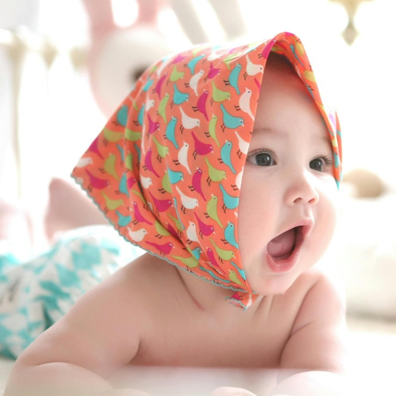 A baby wearing a colorful headscarf, lying on a bed surrounded by soft toys and plush bears. The baby looks curious and joyful, capturing a moment of playful innocence.