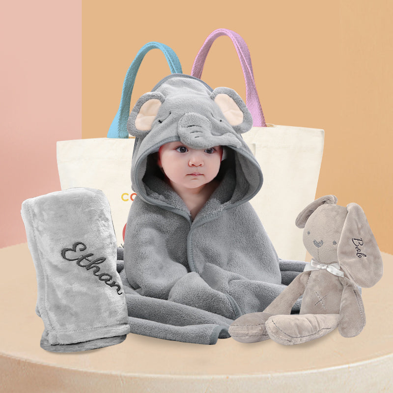A newborn baby wearing a grey elephant hooded bath towel, surrounded by grey soft toys and a grey fleece blanket. The fleece blanket and soft toys feature embroidered names. Displaying a delightful newborn baby hamper.