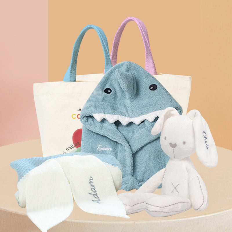 Baby Hamper - Shark Adventure Gift Set. Blue Shark bathrobe, white bunny plushie, soft blue knit blanket with embroidered name on one ear, and Cadeaus brand canvas bag packaging.