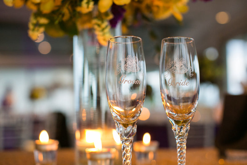 Two champagne glasses labeled 'Bride' and 'Groom' with blurred flowers and candles in the background.