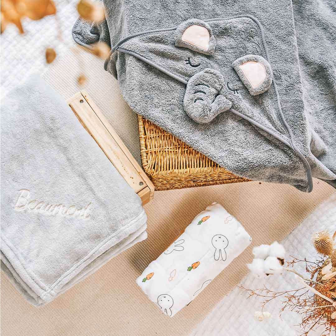 Elephant hooded baby towel, grey with cute embroidered elephant ears and face, displayed with a personalized grey fleece blanket and a Miffy-patterned swaddle.