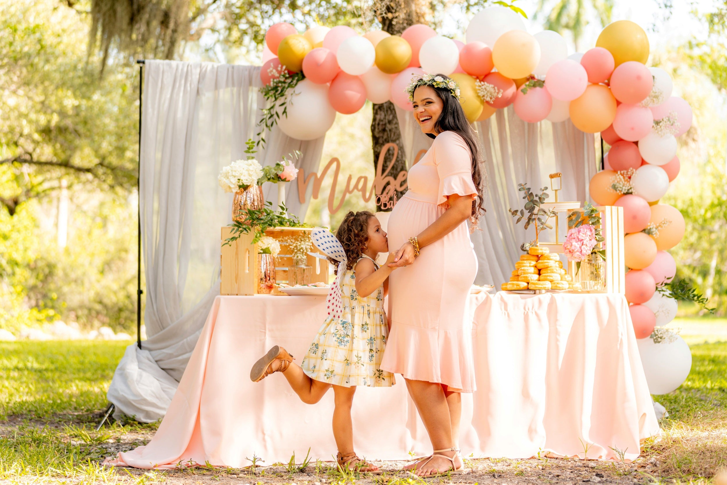 A pregnant woman in a soft pink dress stands next to a beautifully decorated baby shower table, smiling warmly as a young girl kisses her belly. The festive setting features pastel balloons, flowers, and sweet treats, creating a joyful and loving atmosphere perfect for a baby celebration.