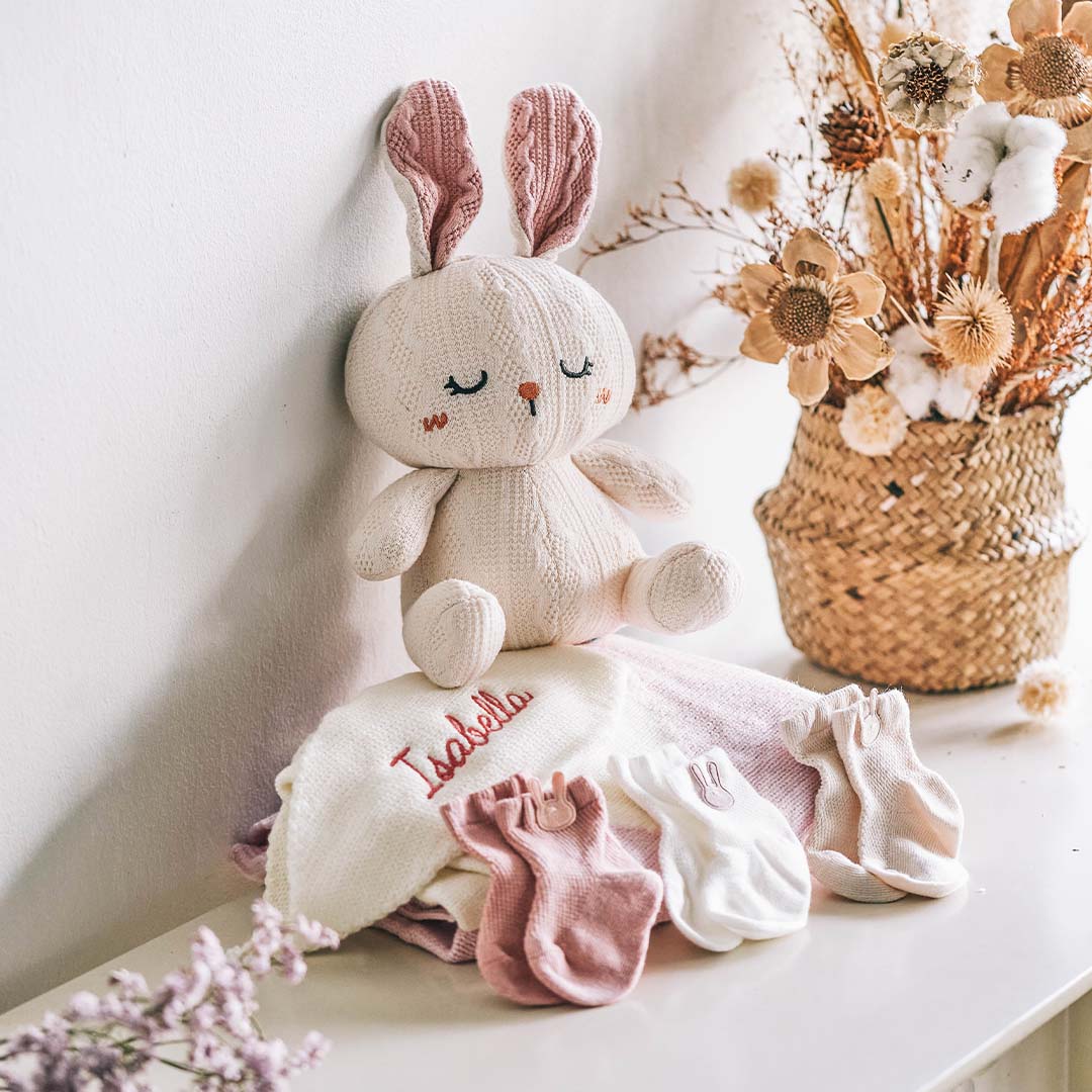 Soft knitted bunny toy sitting beside personalized baby blanket with the name ‘Isabella’ embroidered on it, accompanied by baby socks in pink and white, displayed on a table with a decorative vase of dried flowers.
