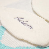 A close-up of a knitted blanket with the name "Adam" embroidered on it. The blanket is light-colored with a garter stitch border.