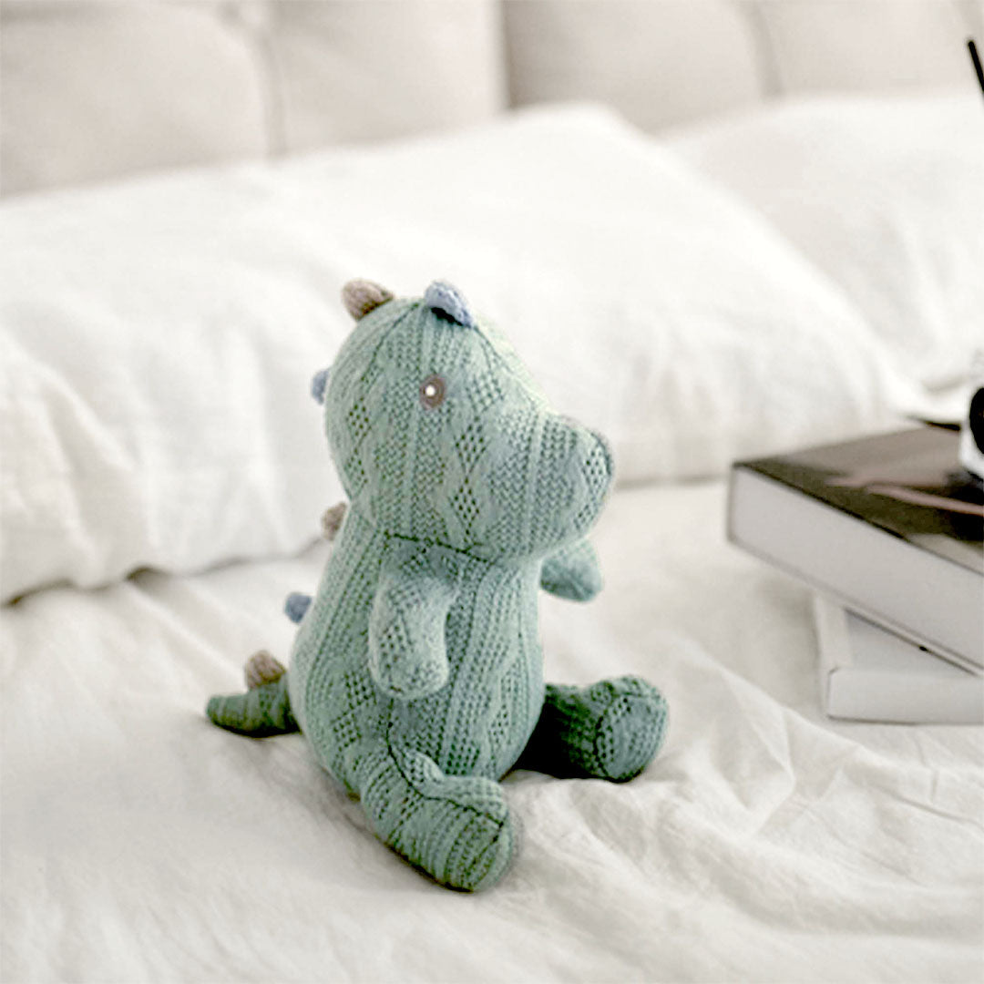 A charming knitted dinosaur toy in a soft seafoam green sits on a white bedspread, gazing out with tiny bead eyes. Its pose is playful and upright, suggesting curiosity and friendliness. The dinosaur is meticulously crafted with a textured knit pattern that gives it a cozy, hand-made feel, ideal for a child&