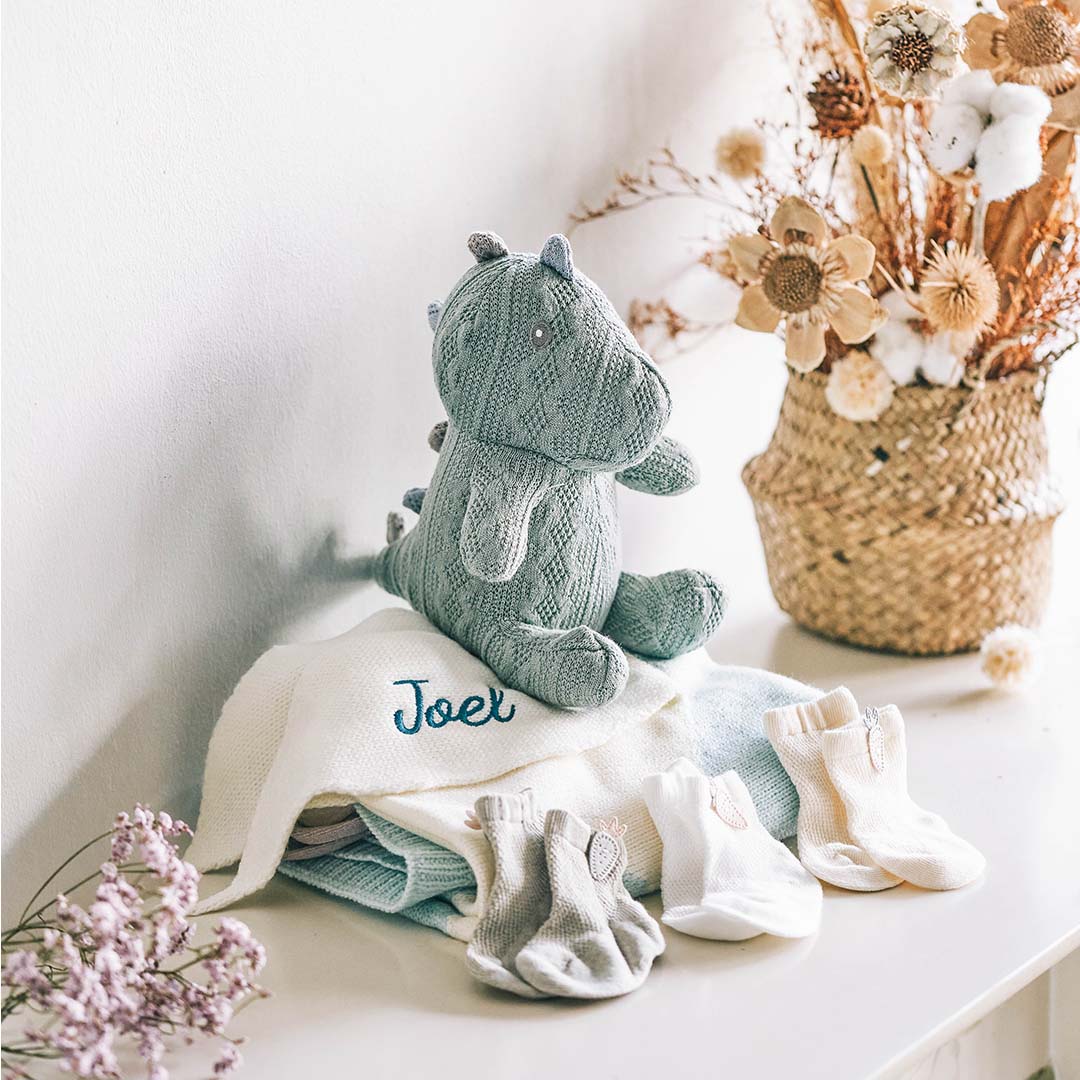 A stuffed dinosaur sitting on a turquoise bunny blanket with 3 pair of socks on a table. There is text on the blanket that says &quot;Joel”.