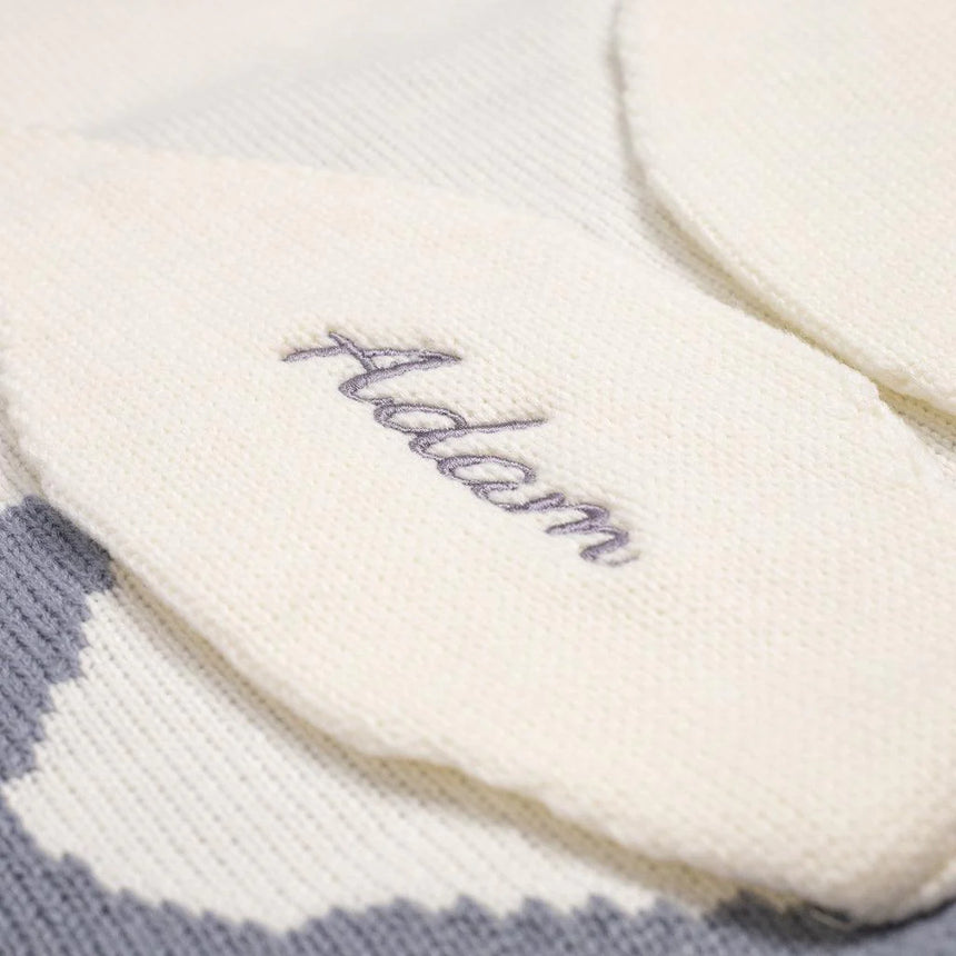 Close-up view of a white knitted baby blanket, showcasing the name 'Adam' elegantly embroidered in gray cursive script on the fabric, emphasizing the personalization detail and the soft texture of the blanket.