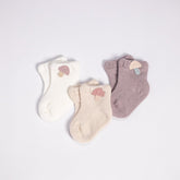 An array of soft, knitted baby socks in a gentle pastel palette, laid out on a white background. The set includes three pairs: one in white, one in beige, and one in mauve, each adorned with a cute knitted patch— an acorn on the white and beige socks and an ice cream on the mauve socks.