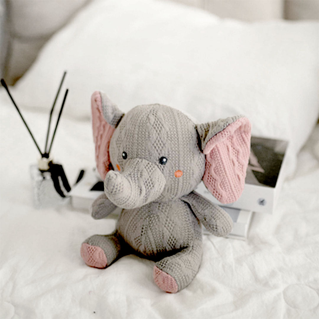 A cozy hand-knitted elephant toy sits on a fluffy white bedspread, exuding a soft and welcoming charm. The elephant, in shades of gray with contrasting pink inner ears and small orange bead eyes, offers a friendly gaze. It&