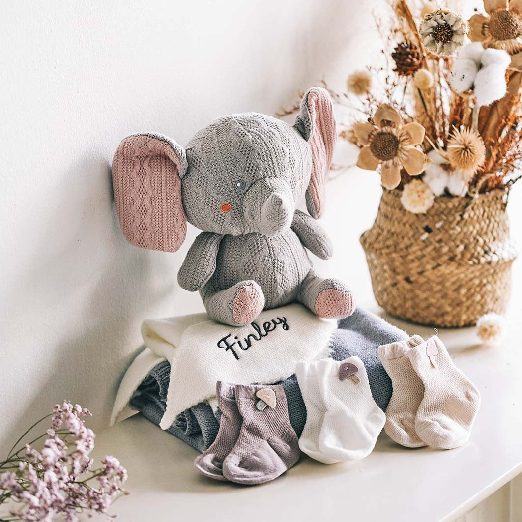 Cozy baby gift set featuring a knitted gray elephant plushie with pink inner ears, sitting atop a stack of soft baby blankets embroidered with the name &