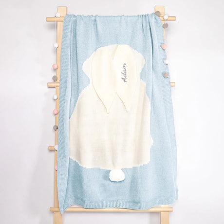 Personalized baby towel draped over a wooden rack, showcasing a delicate pastel blue color with a plush white hood featuring a playful pompom. The hood is elegantly embroidered with the name 'Adam’ in script, set against a clean and simple backdrop that highlights the towel's softness and inviting texture, ideal for a newborn's sensitive skin.