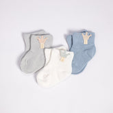 An elegant set of baby socks in soft, pastel tones displayed on a white background. The set includes three pairs: one in light gray, one in soft blue, and one in white, each adorned with a tiny wooden tag featuring a crown emblem. These cozy socks are designed to keep little feet warm, combining functionality with a delicate, stylish look.