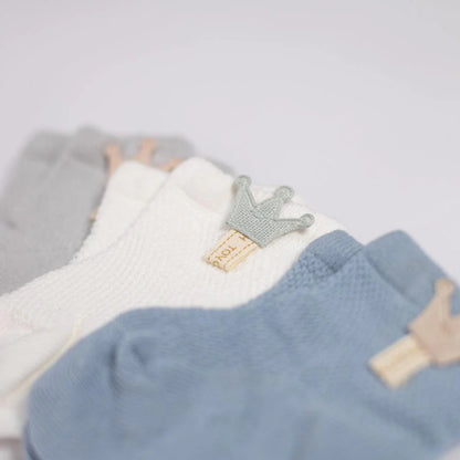 A close-up view of a charming set of baby socks arranged in a fan shape, showcasing intricate textures and muted colors. The collection includes socks in soft gray, pristine white, and serene blue, each topped with a small, textured crown affixed to a beige label, symbolizing a touch of royalty. The soft focus emphasizes the delicate fabric and cozy design, perfect for a newborn.