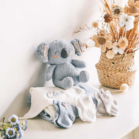 Charming baby gift set on a white shelf, featuring a hand-knitted gray koala plushie with orange button eyes and floppy ears, sitting next to a personalized white baby blanket embroidered with the name 'Cleo'. The set includes three pairs of soft baby socks in shades of gray and white, displayed in front of a rustic woven basket filled with dried flowers, creating a cozy and inviting nursery atmosphere.