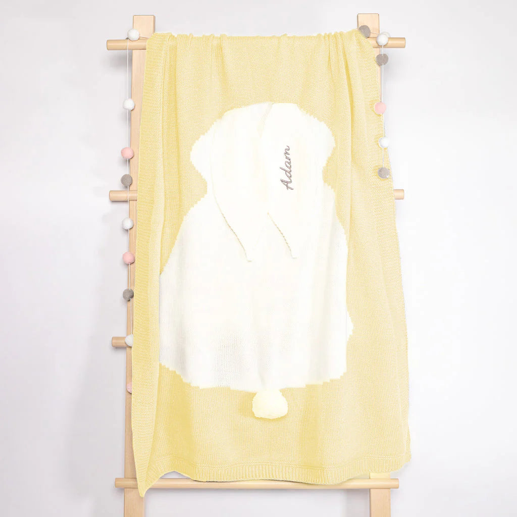 A soft lemon yellow baby towel hangs on a wooden rack, adorned with a plush white hood featuring a cute pompom and elegantly embroidered with the name &