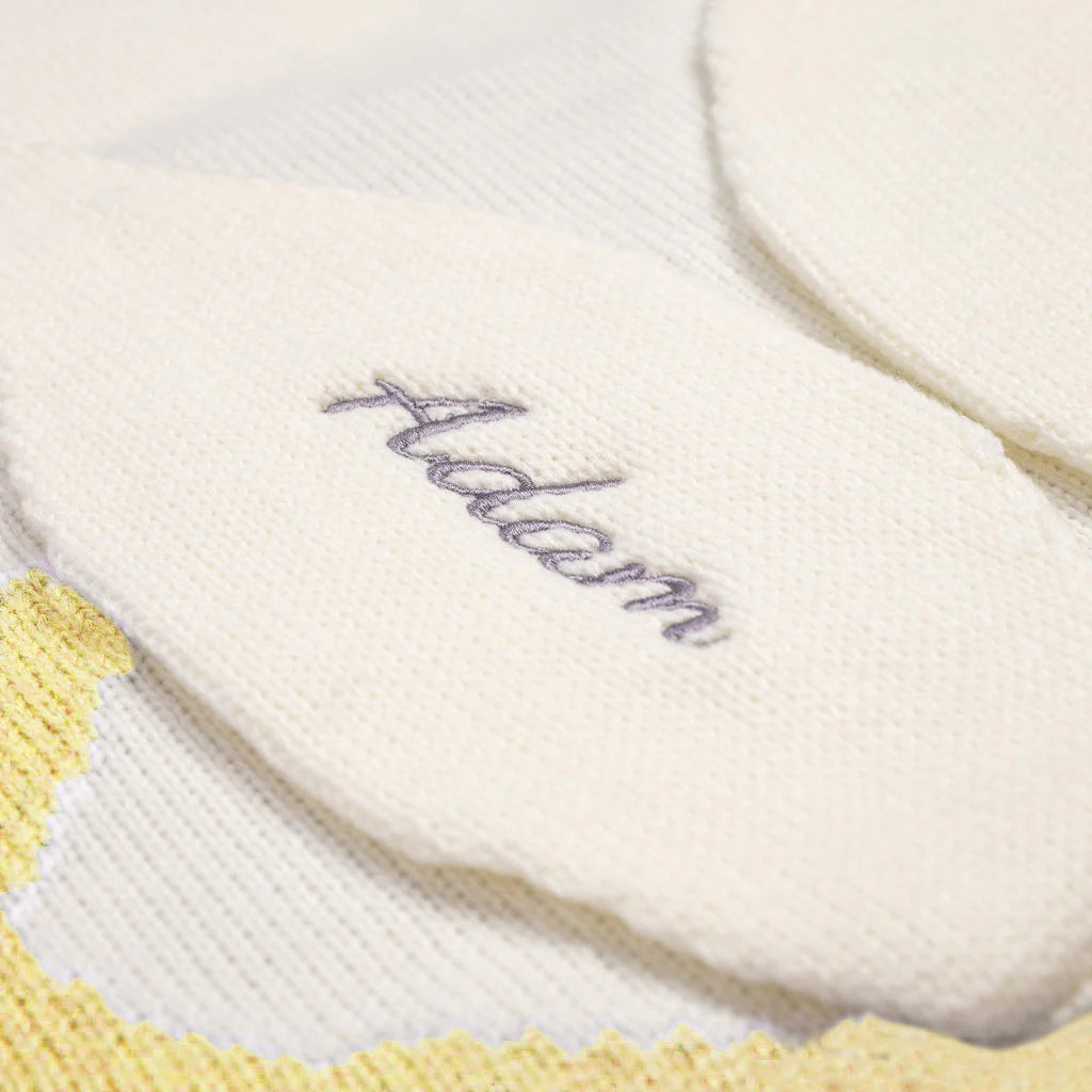 A detailed close-up of a white knitted bunny ear, part of a baby blanket, showcasing intricate embroidery of the name &