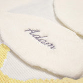 A detailed close-up of a white knitted bunny ear, part of a baby blanket, showcasing intricate embroidery of the name &