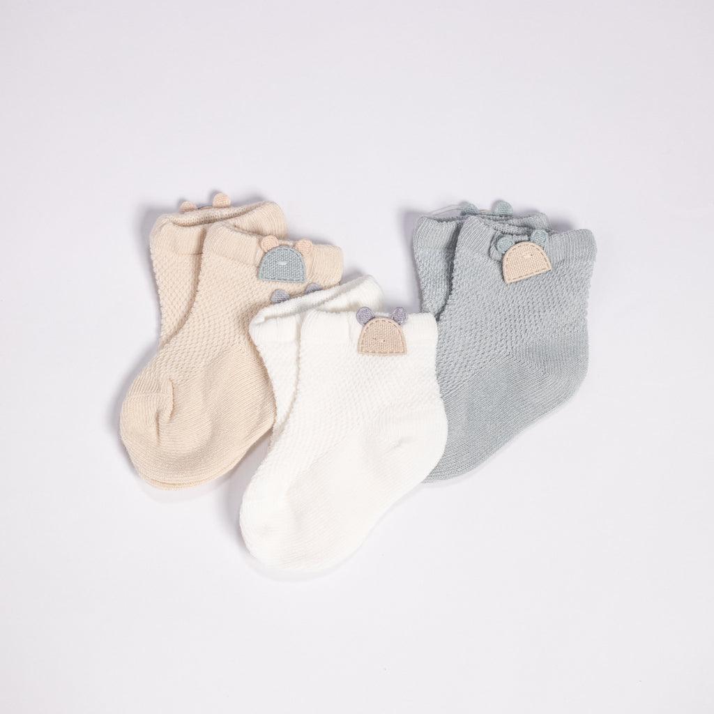 An adorable set of baby socks arranged on a white background. This collection includes three pairs of socks in soft neutral colors: cream, white, and gray. Each sock is decorated with a unique, knitted cuff and adorned with a small, wooden button shaped like a bear&
