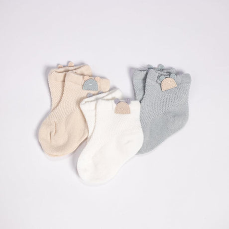 An adorable set of baby socks arranged on a white background. This collection includes three pairs of socks in soft neutral colors: cream, white, and gray. Each sock is decorated with a unique, knitted cuff and adorned with a small, wooden button shaped like a bear's head, adding a whimsical and charming touch to these cozy baby essentials.