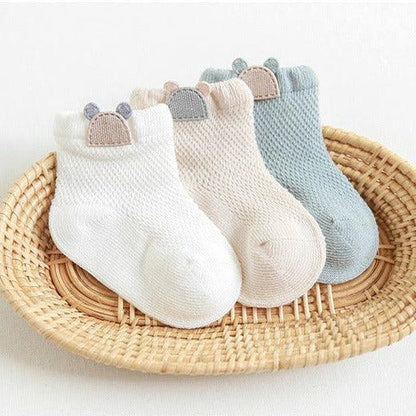 A delightful trio of baby socks presented on a woven rattan tray. These cozy socks are knitted with precision, each in a different pastel hue: crisp white, soft pink, and serene blue. Each pair features a playful ribbed cuff topped with a small, decorative bear-shaped button in muted colors, adding a touch of charm to these essential newborn accessories.