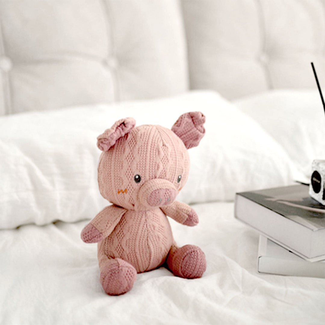 A charming hand-knitted piglet toy in a soft blush pink sits on a white bedspread, bringing a touch of warmth and whimsy to the room. Its playful ears and friendly eyes are accentuated with delicate stitching details, offering a sweet and inviting expression. The piglet&