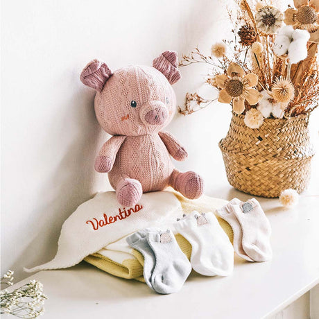 A cozy nursery scene with a hand-knitted pink piglet plush toy sitting proudly on a shelf. The piglet, with its intricate patterns and cheerful blue eyes, accompanies a personalized white blanket embroidered with the name 'Valentina' in a vibrant red thread. Beside them are three pairs of baby socks in neutral colors, each featuring a tiny metallic crown tag. A woven basket filled with dried floral arrangements and cotton bolls adds a rustic charm to the warm and inviting atmosphere.