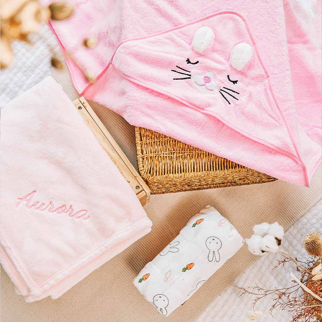 Baby gift set featuring a pink hooded towel with a bunny face, a personalized blanket with ‘Aurora’ embroidered, and a white muslin swaddle with bunny and carrot prints, all arranged next to a wicker basket.