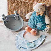 Bear-themed baby dining set featuring a soft bear plush teether with a blue outfit, a BPA-free silicone plate with matching fork and spoon, and a personalized light blue organic cotton bib.