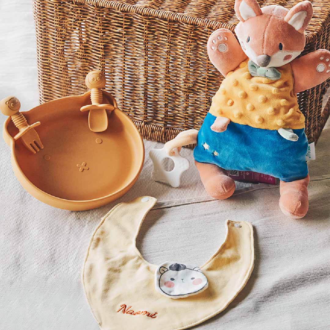 Baby gift set featuring a soft plush fox toy, a personalized bib with ‘Naomi’ embroidered, a silicone baby bowl with matching spoon and fork, and a white star-shaped teether, all arranged in front of a wicker basket.