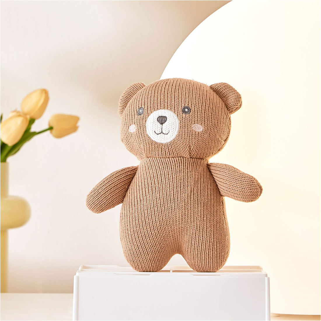 A close-up photo of a brown knitted teddy bear sitting on a white box. The teddy bear has black button eyes, a stitched smile, and a black embroidered nose. It has a small pink patch sewn on its chest.