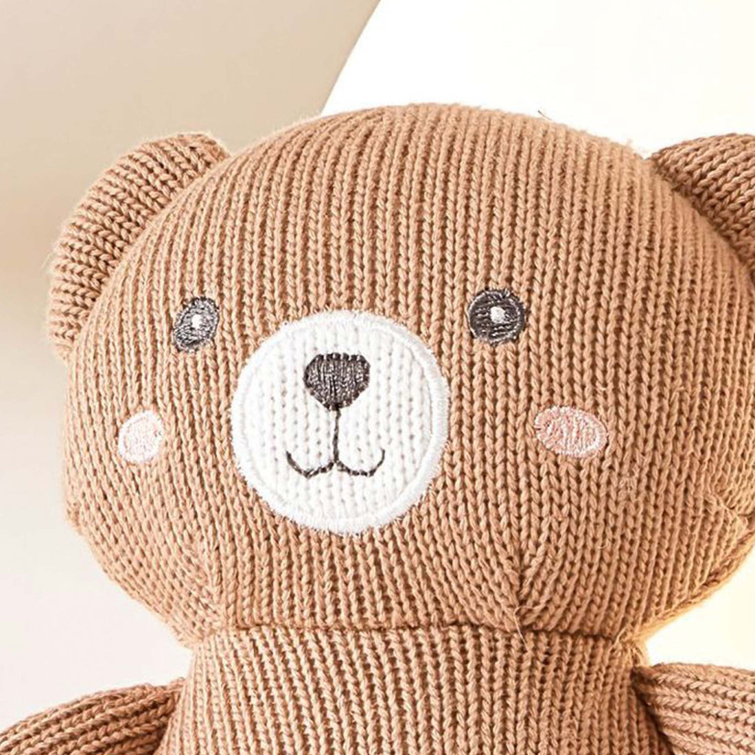 A close-up photo of a brown knitted teddy bear. The teddy bear has black button eyes, a stitched smile, and a black embroidered nose. It has a small pink patch sewn on its chest and is sitting on a white shelf.