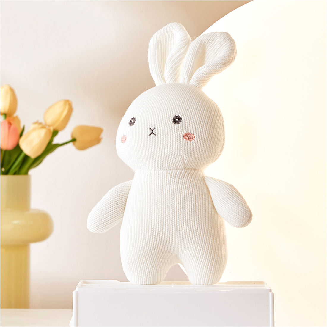 A white stuffed rabbit sitting on a white shelf. The rabbit has floppy ears and a pink cheek.