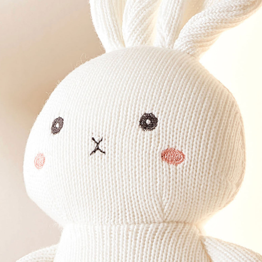A close-up photo of a white knitted rabbit. The rabbit has pink stitching for its eyes, nose, and mouth. It has a pink cheek sewn on its chest.