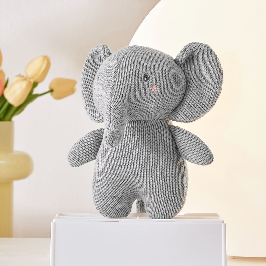 A close-up photo of a stuffed elephant sitting on a white box on a table. The elephant is gray and white with floppy ears and a red bow tie. There is a vase of pink and yellow flowers next to the elephant on the table.