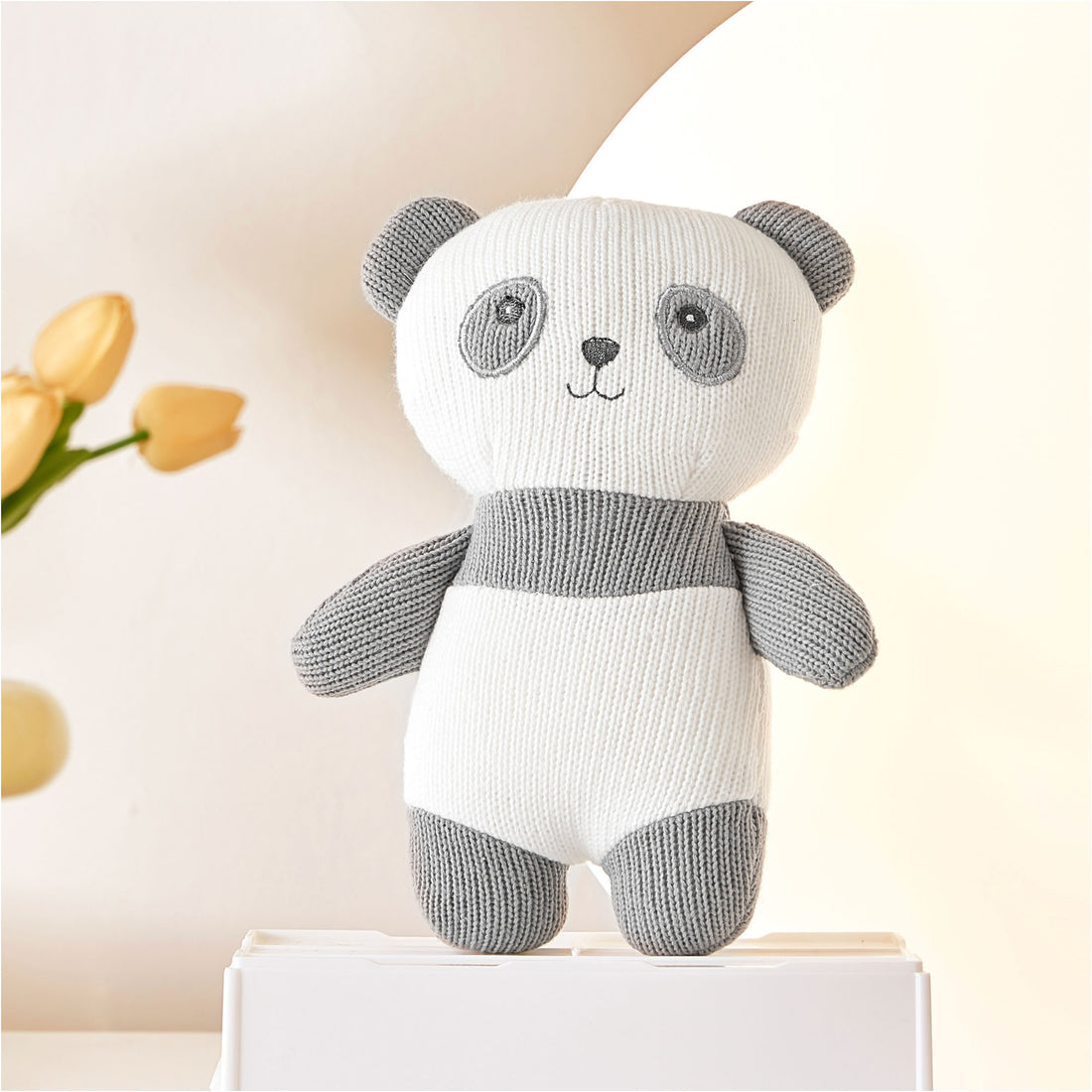 A stuffed panda bear sitting on a white shelf. The panda bear is black and white and made of soft material. It has black eyes, nose, and mouth.