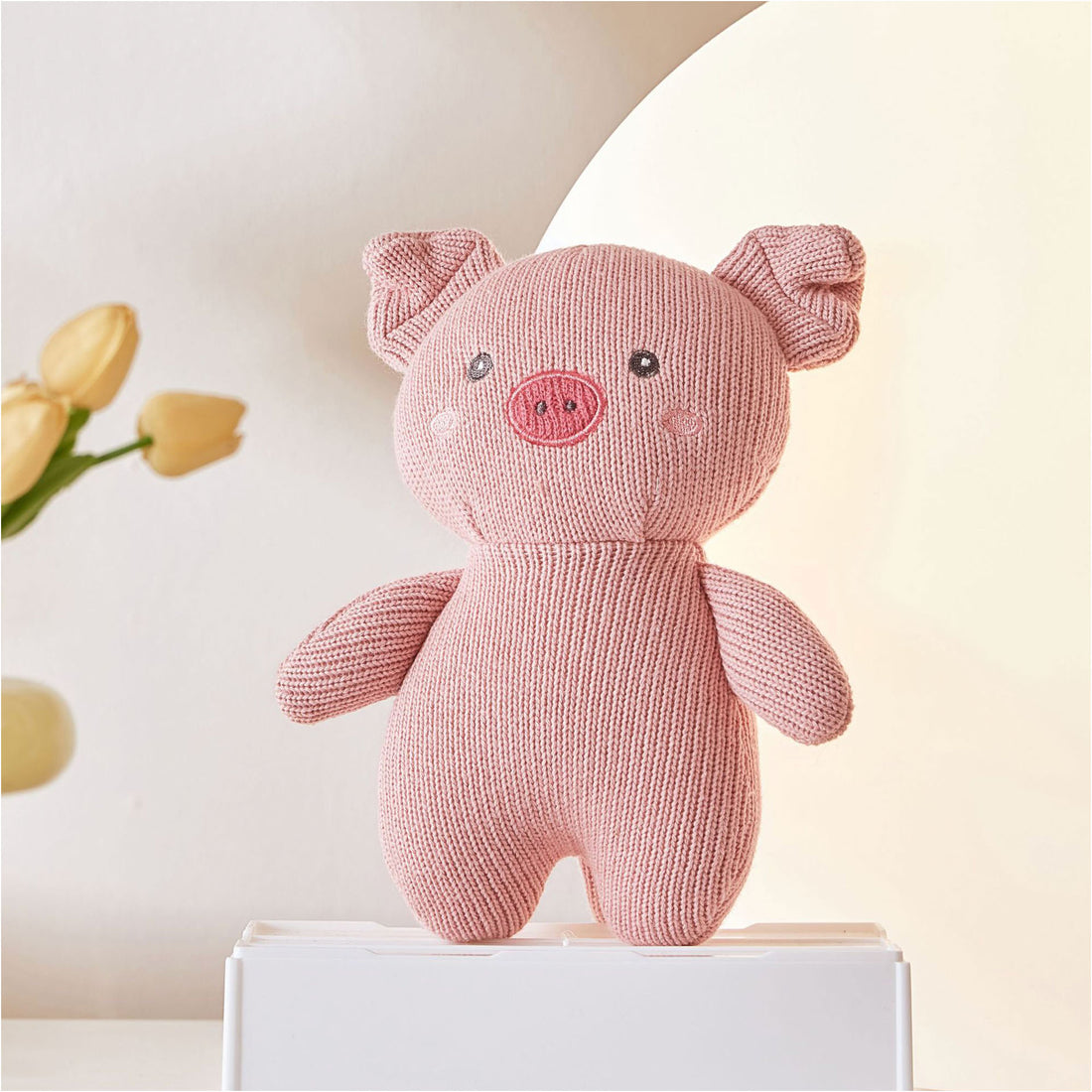 A close-up photo of a pink knitted piggy sitting on a white box. The teddy bear has black button eyes and a stitched smile. It has a black embroidered nose and a small pink patch on its chest.