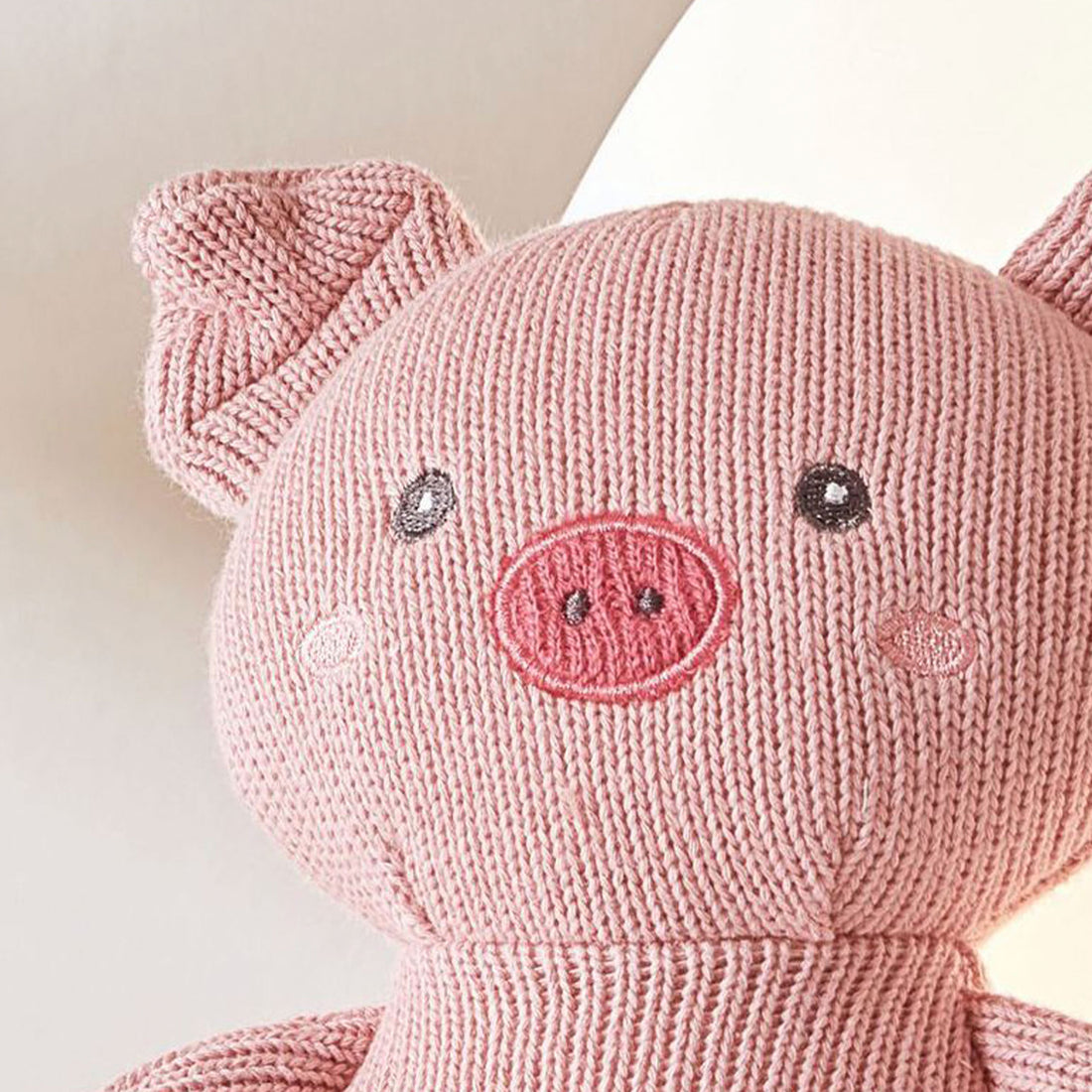 A close-up photo of a pink knitted pig with embroidered eyes and a stitched smile. The pig has a curly tail and small hooves.