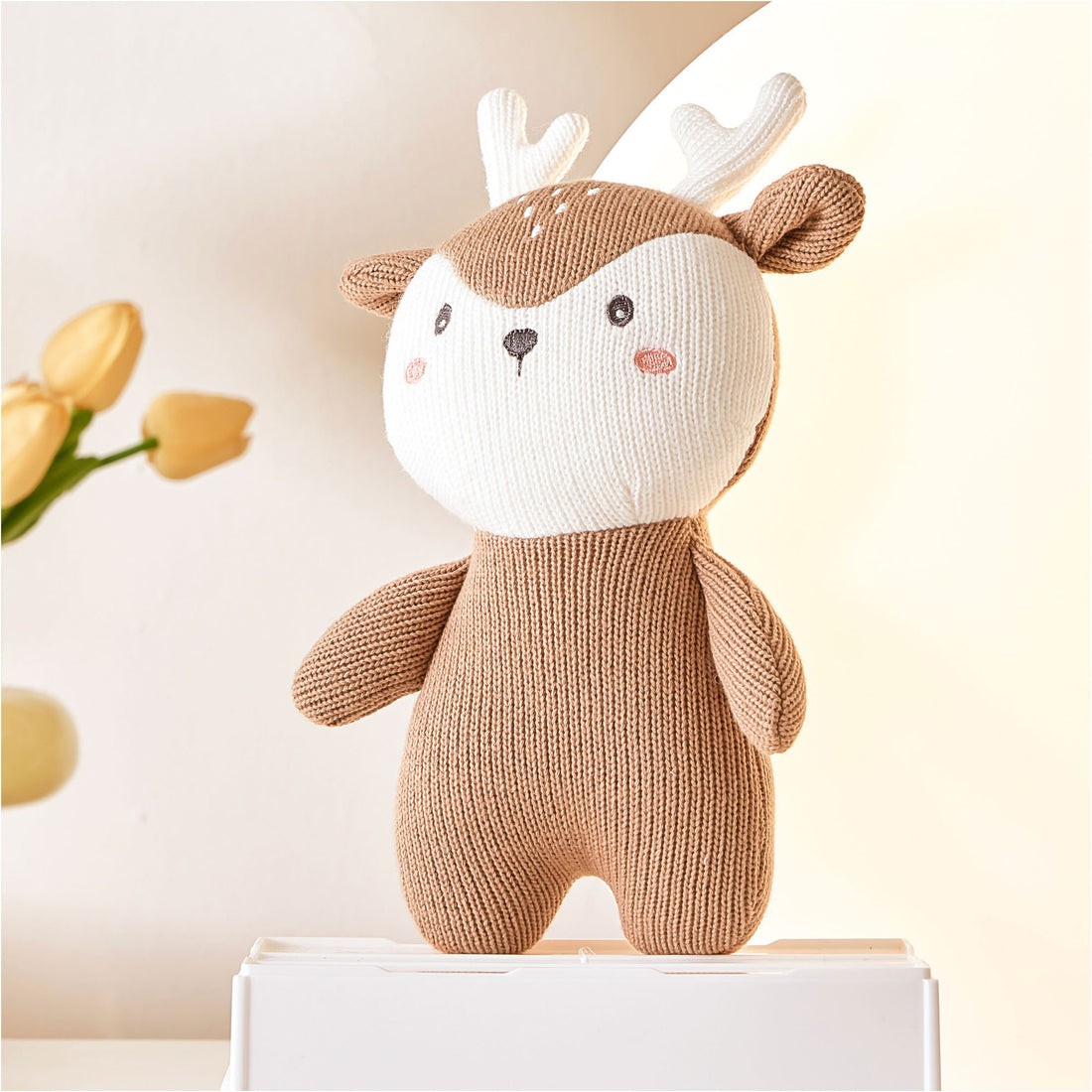 A close-up photo of a stuffed deer sitting on a white shelf. The deer is brown and white with floppy ears and a stitched smile. It has black button eyes and a pink pom-pom for a nose.