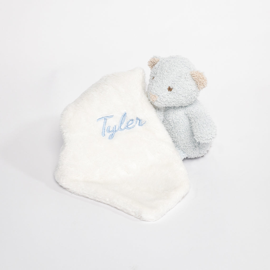 A stuffed animal of a blue bear attached with a white towel with the embroidered name &